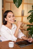 schön asiatisch Mädchen Sitzung im Cafe mit Tasse von Kaffee, Zeichnung auf Digital Tablette mit Grafik Stift, Herumtollen und Herstellung Kratzer foto