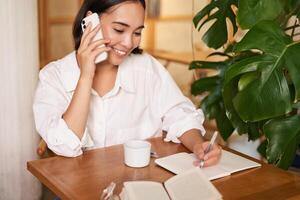 Arbeiten Frau Antworten Telefon Anruf im Cafe, Schreiben runter, Herstellung Anmerkungen während haben Konversation auf Telefon foto