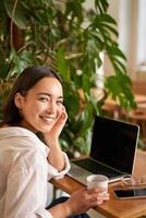 modisch jung Frau Sitzung im Cafe und lächelnd beim Kamera, Trinken Kaffee und mit Laptop, Arbeiten aus der Ferne, studieren oder Surfen Internet foto