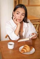 Vertikale Schuss von asiatisch Mädchen sitzt allein im Cafe, liest Buch und sieht aus verärgern, Getränke Kaffee mit Croissant foto