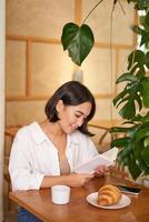 verträumt jung lächelnd asiatisch Frau lesen Buch, Sitzung im Cafe, Essen Croissant und Trinken Kaffee im gemütlich Innere foto