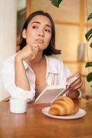 Vertikale Schuss von asiatisch Mädchen sitzt allein im Cafe, liest Buch und sieht aus verärgern, Getränke Kaffee mit Croissant foto