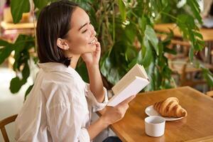 schön jung asiatisch Frau mit ein Buch im Hände, Sitzung im Cafe, Trinken Kaffee und Essen Croissant, lächelnd, suchen mysteriös foto