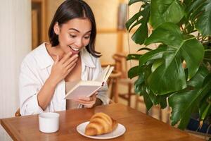 asiatisch Mädchen sieht aus erstaunt beim Seiten, lesen mit Aufregung, Sitzung im Cafe und Essen Croissant foto