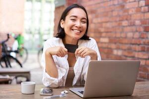 glücklich jung asiatisch weiblich, Sitzung mit Laptop und zeigen Anerkennung Karte, bezahlen Rechnungen online, Einkaufen im Internet, Sitzung im Cafe mit Kaffee foto