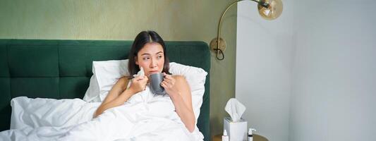 Gesundheit und Personen. asiatisch Mädchen Lügen im Bett, Gefühl krank, Niesen und Trinken heiß Tee, fangen kalt, bleiben beim Zuhause auf verlassen foto