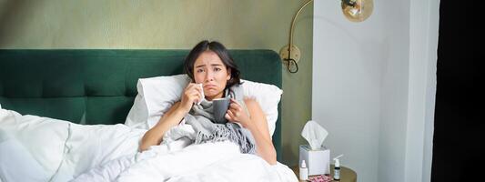 Gesundheit und Personen. asiatisch Mädchen Lügen im Bett, Gefühl krank, Niesen und Trinken heiß Tee, fangen kalt, bleiben beim Zuhause auf verlassen foto