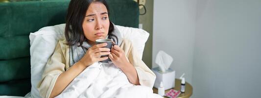 COVID-19, Grippe und Impfung Konzept. Koreanisch Mädchen Lügen im Bett mit kalt, fangen Grippe, Trinken heiß Tee und mit vorgeschrieben Medikation foto