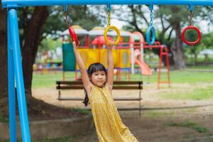 glücklich Mädchen hängend auf Affe Bar durch Hand tun Übung. wenig asiatisch Mädchen spielen beim draussen Spielplatz im das Park auf Sommer- Urlaub. gesund Aktivität. foto