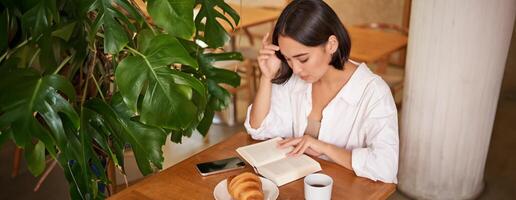 schön jung asiatisch Frau Sitzung im Cafe mit ein Buch, Essen Croissant und Lektüre, Trinken Tasse von Kaffee foto