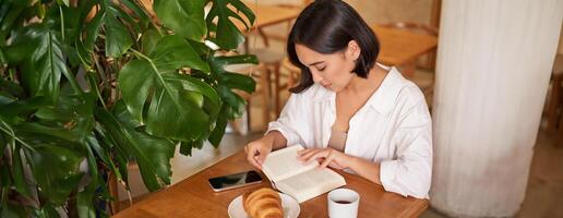 schön jung asiatisch Frau Sitzung im Cafe mit ein Buch, Essen Croissant und Lektüre, Trinken Tasse von Kaffee foto