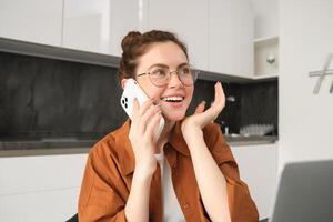 Porträt von jung Geschäftsfrau, Selbstständiger Dame Arbeiten von heim, Herstellung Telefon Anrufe zu Kunden und lächelnd, mit Laptop im Küche foto