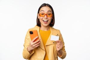 online Einkaufen. stilvoll asiatisch weiblich Modell- im Sonnenbrille, halten Anerkennung Karte und Handy, Mobiltelefon Telefon, lächelnd Glücklich, Stehen Über Weiß Hintergrund foto