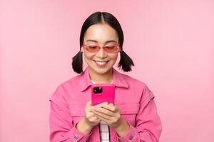 Porträt von asiatisch Mädchen im Sonnenbrille mit Smartphone. Frau suchen beim Handy, Mobiltelefon Telefon, Surfen im Anwendung, Stehen Über Rosa Hintergrund foto