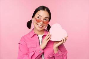 Bild von stilvoll asiatisch Freundin im Sonnenbrille, raten was ist Innerhalb Geschenk Kasten, herzförmig gegenwärtig, Stehen Über Rosa Hintergrund foto