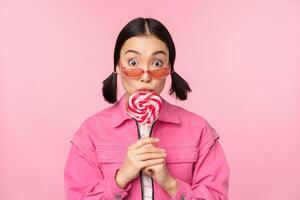 dumm und süß asiatisch weiblich Modell- Lecken lolipop, Essen Süßigkeiten Süss und lächelnd, suchen aufgeregt, Stehen Über Rosa Hintergrund foto