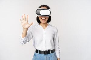 lächelnd asiatisch Frau winken beim Du, tragen virtuell Wirklichkeit Gläser, vr Headset, chatten im Meta Universum, Stehen Über Weiß Hintergrund foto