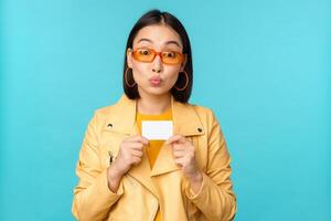 stilvoll jung asiatisch Frau im Sonnenbrille, zeigen Anerkennung Karte und lächelnd, empfehlen Bank, kontaktlos Zahlung oder Rabatte im speichern, Stehen Über Blau Hintergrund foto