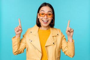 begeistert asiatisch Mädchen im Sonnenbrille, Punkte Finger hoch, zeigt an Banner oder Logo auf Spitze, steht Über Blau Hintergrund foto