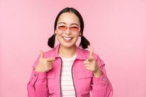 schließen oben Porträt von modern asiatisch Mädchen im Sonnenbrille lächelnd, zeigen Finger beim Kamera, loben Du, einladend oder Kompliment machen, Stehen Über Rosa Hintergrund foto