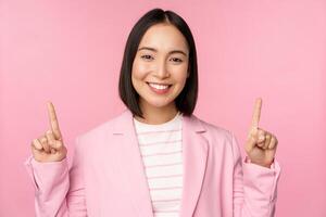 Porträt von asiatisch Geschäftsfrau zeigen Finger oben und lächelnd, zeigen Geschäft Unternehmen Logo, Information auf Spitze, Stehen Über Rosa Hintergrund foto