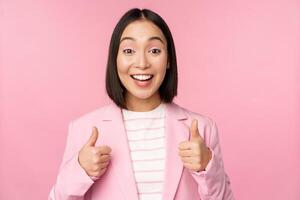 Porträt von asiatisch Geschäftsfrau lächelnd befriedigt, zeigen Daumen hoch, loben, mögen und genehmigen, Stehen im passen Über Rosa Hintergrund foto