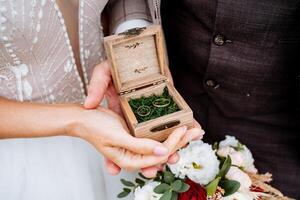 das Bräutigam im das passen und das Braut im ein Schneewittchen elegant Kleid halt ein hölzern Box mit Hochzeit Ringe. im das andere Hand, das Braut hat ein Hochzeit Strauß von Weiß und rot Blumen. foto