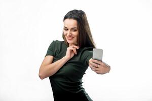 froh jung Frauen Herstellung Selfie durch ihr Smartphone. glücklich ziemlich Mädchen mit Gerade dunkel Haar macht Selfie mit Fantastisch Lächeln gekleidet schwarz T-Shirt auf das Hintergrund von Weiß Mauer. foto