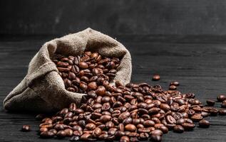 organisch Kaffee Saat Streuung von das Sack Über das dunkel hölzern Oberfläche. frisch Kaffee Bohnen in der Nähe von das Licht braun Sack isoliert. Nahansicht foto