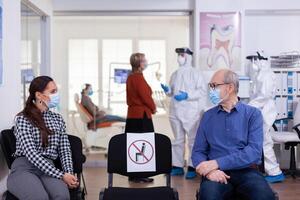 Senior Mann mit Gesicht Maske diskutieren mit Frau geduldig im Stomatologie Klinik im warten Zimmer, behalten Sozial Distanzierung während global Pandemie mit Coronavirus. Arzt Stehen tragen ppe Anzug. foto