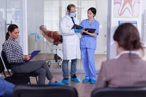 Zahnarzt zeigen Zähne Röntgen Überprüfung es mit Krankenschwester Arzt und Assistent Arbeiten im modern überfüllt stomatologisch Klinik, Patienten Sitzung auf Stühle im Rezeption Füllung im Dental Formen warten. foto