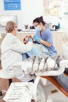 Stomatologe Senior Arzt tun Zahnschmerzen Behandlung auf krank geduldig während Stomatologie Beratung. Mann Sitzung auf kieferorthopädisch Stuhl im Krankenhaus Büro während medizinisch Krankenschwester Reinigung Teeh foto