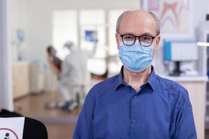 Porträt von Alten Mann im Dental Büro suchen auf Kamera tragen Gesicht Maske Sitzung auf Stuhl im warten Zimmer stomatologisch Klinik. Konzept von Neu normal Zahnarzt Besuch im Coronavirus Ausbruch. foto