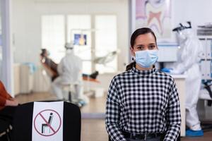 Porträt von Frau im Dental Büro suchen auf Kamera tragen Gesicht Maske Sitzung auf Stuhl im warten Zimmer Klinik während Arzt Arbeiten. Konzept von Neu normal Zahnarzt Besuch im Coronavirus Ausbruch. foto