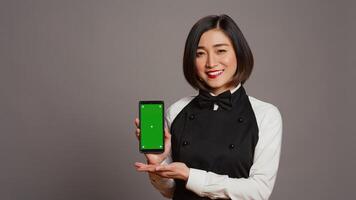 Restaurant Hostess präsentieren Smartphone mit grüner Bildschirm, zeigen isoliert Anzeige mit Chromakey Layout im Studio. asiatisch Kellnerin mit Schürze halten Telefon mit Exemplar. Kamera b. foto
