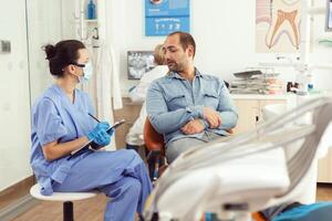 Kieferorthopäde Krankenschwester diskutieren mit Mann geduldig Über Zahn Beratung Schreiben Gesundheitswesen Behandlung auf Zwischenablage Sitzung im medizinisch Dental Büro. Stomatologe Herstellung Chirurgie geplanter Termin foto