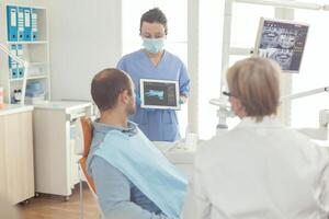 Stomatologe Krankenschwester zeigen Zahn Röntgen zu krank geduldig erklären Behandlung mit Tablette Arbeiten im Stomatologie Krankenhaus Büro. Senior Arzt Prüfung Zahnschmerzen während Mann Sitzung auf Dental Stuhl foto