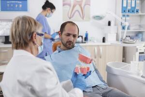 Senior Frau Arzt zeigen künstlich Kiefer Modell- zu geduldig mit Zahnschmerzen diskutieren Träger Behandlung während Stomatologie Beratung im Zahnheilkunde Büro Zimmer. Stomatologe erklären Oral Hygiene foto