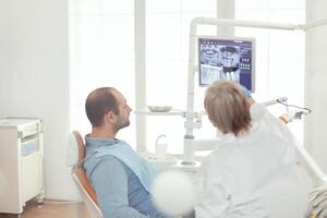 krank Mann Sitzung auf Dental Stuhl suchen beim Zähne Radiographie vorbereiten zum Somatologie Operation. Senior Arzt erklären Zahnschmerzen Schmerzen vorbereiten medizinisch Behandlung während Arbeiten im kieferorthopädisch Büro foto