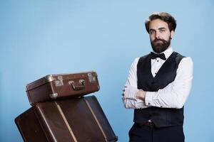 formal Hotel Arbeiter posieren mit Gepäck, tragen formal passen und Weiß Handschuhe Über Blau Hintergrund. männlich Hotelpage Stehen Nächster zu Stapel von tolly Taschen und Koffer, Gastfreundschaft Industrie. foto