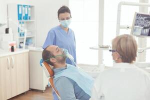 krank geduldig Sitzung auf Dental Stuhl tragen Oxigen Maske während Stomatologie Termin. Zahnärzte Krankenhaus Mannschaft Prüfung Zahn während Arbeiten im Zahnheilkunde Kabinett vorbereiten Gesundheitswesen Behandlung foto