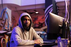 afrikanisch amerikanisch Hacker tun illegal Aktivitäten auf Computer und Arbeiten spät Porträt. kriminell im Kapuze brechen Webseite mit ddos Attacke und suchen beim Kamera im verlassen Warenhaus foto