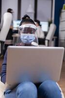 afrikanisch Geschäftsfrau Arbeiten auf Laptop tragen Gesicht Maske gegen COVID-19. multiethnisch Geschäft Mannschaft Arbeiten respektieren Sozial Entfernung während global Pandemie mit Coronavirus. foto