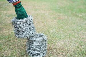 schließen oben Arbeiter Hand tragen Grün Handschuhe, hält Rollen von mit Widerhaken Kabel. Konzept, Konstruktion Werkzeug. mit Widerhaken Draht ist benutzt zum machen Zäune , sichern Eigentum ,machen Rand zu Show das Gebiet von Bereich. foto