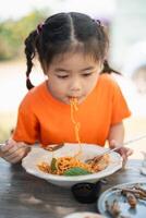 jung Mädchen Kinder genießen ein gabelreich von Spaghetti. Nahansicht von ein wenig Mädchen im ein Orange Hemd Essen Spaghetti mit ein konzentriert Ausdruck, draußen Restaurant. foto