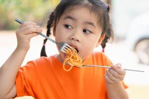 jung Mädchen Kinder genießen ein gabelreich von Spaghetti. Nahansicht von ein wenig Mädchen im ein Orange Hemd Essen Spaghetti mit ein konzentriert Ausdruck, draußen Restaurant. foto