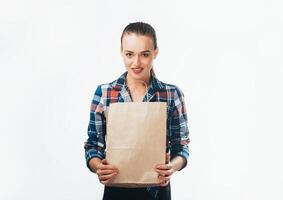 ein lächelnd Frau mit ein Papier Tasche im ihr Hände. ein schön Mädchen im ein Plaid Hemd halten ein Einkaufen Tasche auf Weiß Hintergrund. foto