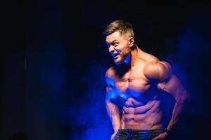 stark brutal Bodybuilder Mann mit perfekt Abs, Schultern, Bizeps, Trizeps und Truhe zeigen seine Bizeps. Schrei Motivation. Blau Filter auf dunkel Hintergrund foto