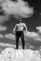 einfarbig Foto von sportlich Bodybuilder posieren. stark attraktiv Mann Stehen auf Weiß Berg mit schön Aussicht von Himmel. männlich halten Mode Sonnenbrille.