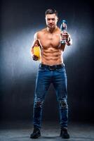 mit nacktem Oberkörper sportlich muskulös Kerl halten Flaschen mit immer noch Getränke im Hände auf das dunkel grau bachground. voll Größe Foto. Porträt. Nahansicht foto